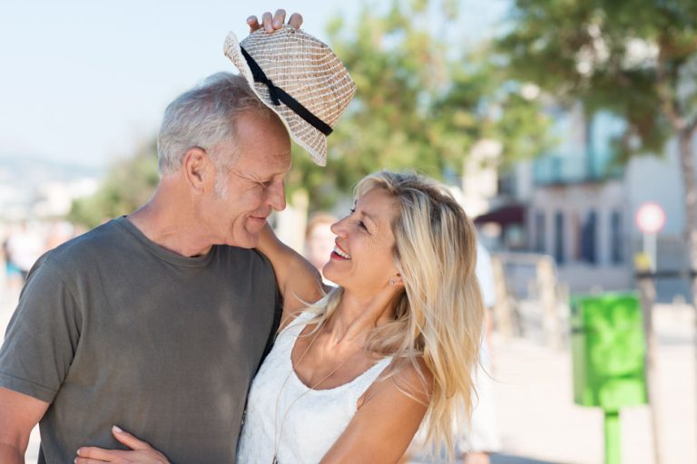Laughing beautiful affectionate woman giving her husband her trendy hat as they enjoy a summer vacation at the coast